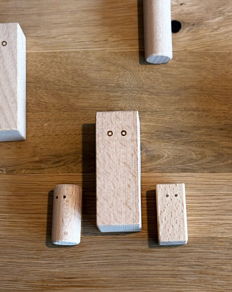 verschieden große Holzklötzchen, teils Rund, teils eckig liegen auf einem Holzuntergrund. Sie haben zwei kleine Kreise, die aussehen wie Augen. Sie sind ein Arbeitsmittel