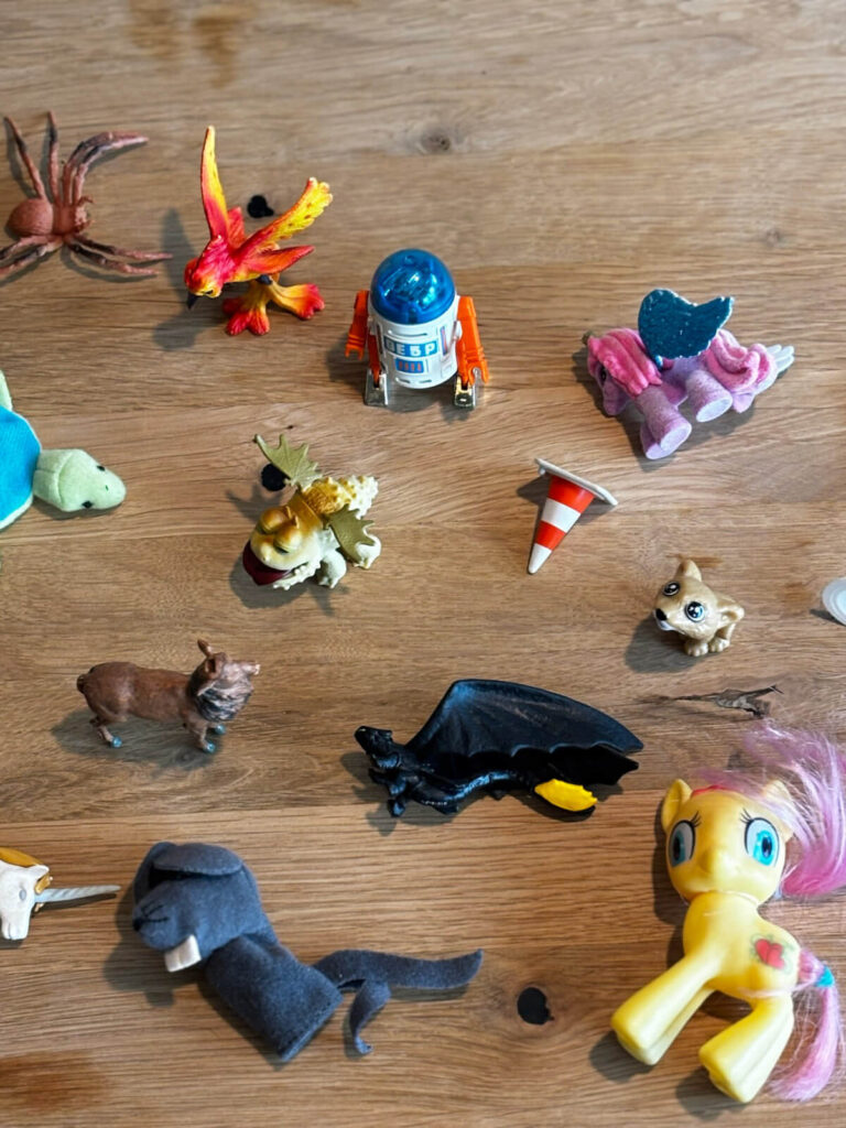 Eine Sammlung verschiedener Spielzeugfiguren, wie ein Einhorn, Drache, R2D2 oder ein Reh. Sie liegen auf einem Untergrund aus warmen Holz und symbolisieren eine Arbeitsmethode